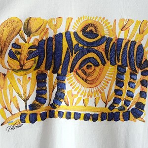 Op de afbeelding: Een wit T-shirt met een gele en blauwe illustratie van een tijger in een bloemenveld. De tijger heeft blauwe strepen en de bloemen zijn geel. De tekst "Ukraine" is in blauw en geel gedrukt aan de onderkant van het T-shirt.