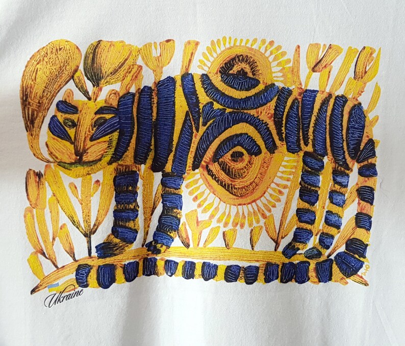 Op de afbeelding: Een wit T-shirt met een gele en blauwe illustratie van een tijger in een bloemenveld. De tijger heeft blauwe strepen en de bloemen zijn geel. De tekst "Ukraine" is in blauw en geel gedrukt aan de onderkant van het T-shirt.