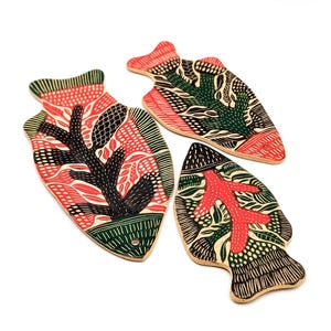 May include: Three wooden, fish-shaped decorative pieces with intricate patterns. Each piece features a unique design in red, green, black, and white. The designs include floral and geometric elements, creating a handcrafted aesthetic.
