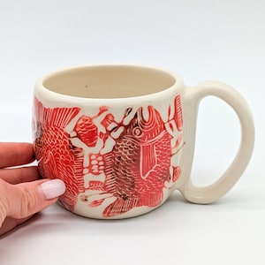 Può includere: Una tazza in ceramica bianca con un design di pesci rossi e arancioni. La tazza ha un manico ed è perfetta per contenere bevande calde.