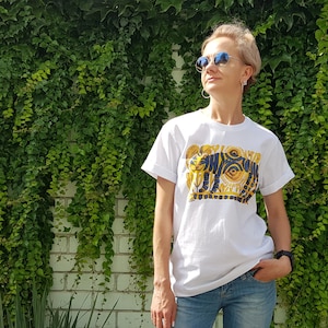 Op de afbeelding: Een wit T-shirt met een grafische print van een gestileerde tijger met een groot oog en zon-achtige ontwerpen in geel en blauw.