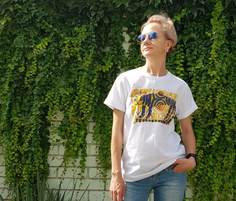 Op de afbeelding: Een wit T-shirt met een grafische print van een gestileerde tijger met een groot oog en zon-achtige ontwerpen in geel en blauw.