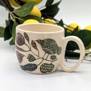 Może przedstawiać: Kremowy kubek ceramiczny z ręcznie rysowaną ilustracją ptaków i liści w odcieniach ciemnej zieleni i brązu. Kubek ma zaokrąglony korpus, szeroki otwór oraz zakrzywione ucho po prawej stronie, wykończone gładką, błyszczącą glazurą.