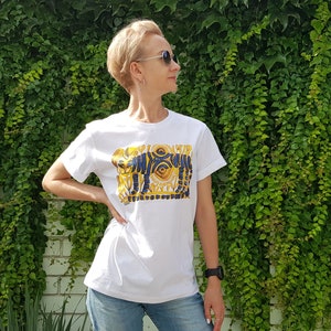 Op de afbeelding: Wit T-shirt met een grafische print van een gestileerd ontwerp in blauw en goud met een oog en geometrische vormen.
