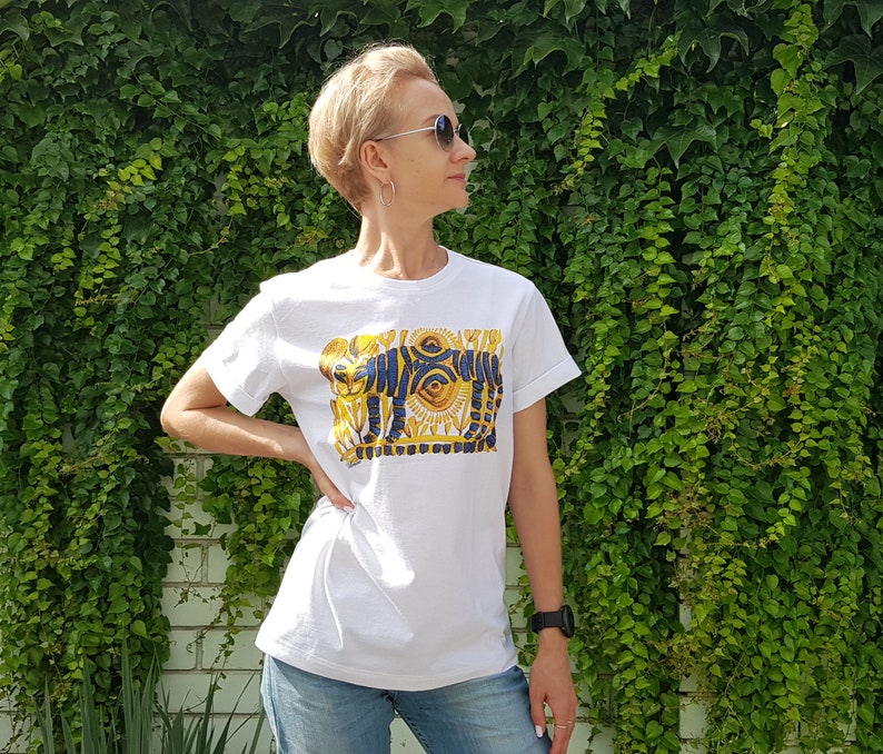 Op de afbeelding: Wit T-shirt met een grafische print van een gestileerd ontwerp in blauw en goud met een oog en geometrische vormen.