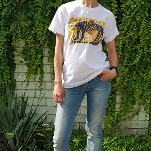 Op de afbeelding: Een wit T-shirt met een blauwe en gouden afbeelding van een tijger met een zon en ogen. De persoon die het shirt draagt staat voor een bakstenen muur met groene klimplanten.