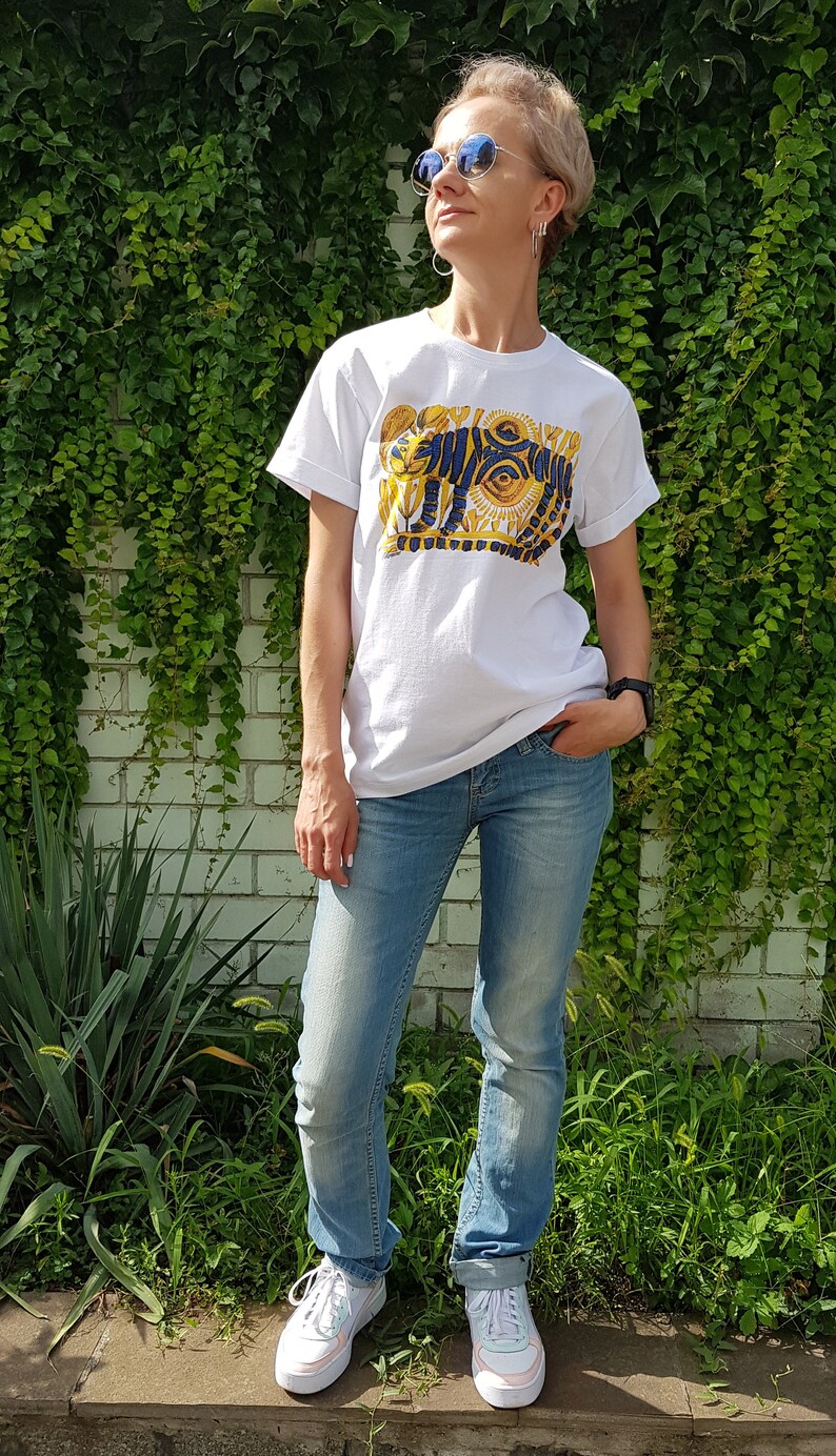 Op de afbeelding: Een wit T-shirt met een blauwe en gouden afbeelding van een tijger met een zon en ogen. De persoon die het shirt draagt staat voor een bakstenen muur met groene klimplanten.