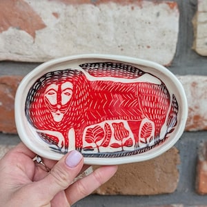 Op de afbeelding: Een witte keramische schaal met een rood en zwart leeuwenontwerp. De leeuw is afgebeeld in een gestileerde folk-art stijl met een gedetailleerde manen en een zwarte omtrek.