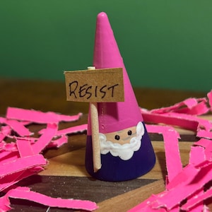 May include: A whimsical gnome figurine with a pink hat and a blue body, holding a sign that reads "RESIST." The gnome has a white beard and is surrounded by pink paper strips. The figurine is on a wooden surface.