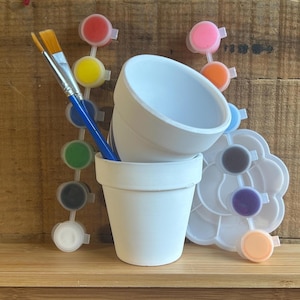 May include: Two white ceramic flower pots, a paintbrush, and a paint palette with various colors. The paint palette is white and shaped like a flower. The paintbrushes have blue handles and yellow bristles. The pots are empty.