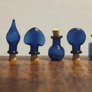 May include: Six cobalt blue glass bottle stoppers with various shapes, including a sphere, teardrop, and mushroom design. Each stopper has a black base and a natural cork. The stoppers are arranged on a wooden surface.