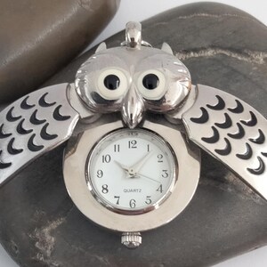 Op de afbeelding: Zilverkleurig zakhorloge in de vorm van een uil met een witte wijzerplaat. De vleugels van de uil zijn uitgespreid en de wijzerplaat geeft de tijd weer. Het horloge heeft een quartz uurwerk en een lus om een ketting aan te bevestigen.