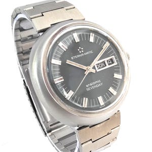 Puede incluir: Un reloj de pulsera vintage Eterna-Matic anatomic SEVENDAY con una correa y caja de acero inoxidable plateado. La esfera del reloj es gris oscuro con agujas e indicadores plateados. El día y la fecha se muestran a las 3 en punto. Un reloj clásico.
