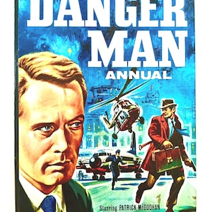 May include: Vintage "Danger Man Annual" book cover. The cover features a portrait of a man with blue eyes and the title "Danger Man Annual" in large white letters. The cover also shows a helicopter, a plane, and a man in a red coat.