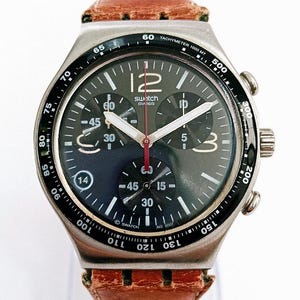 May include: A Swatch chronograph wristwatch with a dark face, silver-tone hands, and a red second hand. The dial features three sub-dials, a date window at the four o'clock position, and a black tachymeter bezel. It has a brown leather strap.