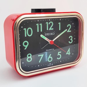 Puede incluir: Un reloj despertador Seiko de cuarzo rojo vintage con esfera negra y borde dorado. El reloj presenta números verdes, un segundero rojo y el logotipo de Seiko. El reloj está marcado "Japan" en la parte inferior.
