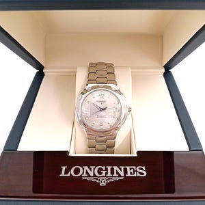 May include: A silver-tone Longines Conquest Automatic wristwatch with a metallic link bracelet, featuring a round silver dial with Arabic numerals at 6 and 12 o'clock, a date window at the 3 o'clock position, and the Longines logo on the watch face.