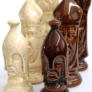 Puede incluir: Un conjunto de piezas de ajedrez de cerámica en tonos marrón y crema. Cada pieza presenta un diseño detallado de rostro. Las piezas están dispuestas sobre un fondo blanco, mostrando sus formas y texturas únicas.