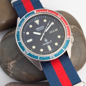 Puede incluir: Un reloj Seiko Quartz SilverWave Z con esfera negra, marcadores luminosos y visualización de la fecha. El reloj tiene un bisel giratorio con detalles en rojo y azul y una correa de nailon a rayas azules, rojas y blancas.