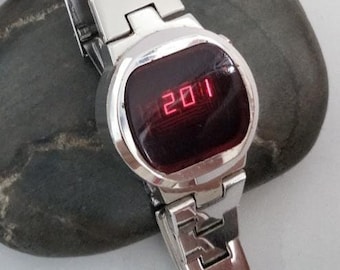May include: A silver digital watch showing the number "2011" in red on a dark display. The watch has a rectangular face with rounded corners and a linked metal strap. The watch is on a dark grey stone.