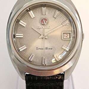 Puede incluir: Reloj Rado Space Wing plateado con esfera gris claro, que muestra la fecha 12 en un recuadro rojo. El reloj tiene marcadores de hora y agujas plateadas, y una correa de cuero negro.