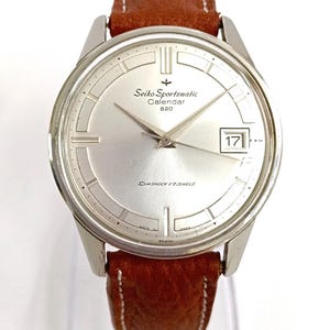 Peut inclure: Une montre-bracelet Seiko Sportsmatic Calendar 820 avec un cadran argenté, des aiguilles argentées et un guichet de date affichant 17. La montre présente un bracelet en cuir marron texturé avec des coutures blanches et un boîtier en métal argenté.