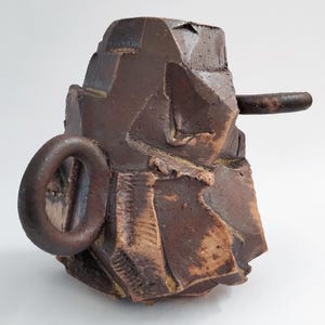 Puede incluir: Escultura de cerámica abstracta hecha a mano en tonos marrones y beige. La pieza presenta una forma geométrica y angulosa con un asa circular y una varilla horizontal. La textura rugosa y los tonos terrosos sugieren una estética rústica.