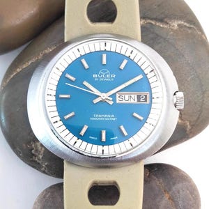 Könnte beinhalten: Eine Vintage Buler Tasmania Uhr mit blauem Zifferblatt, silberner Lünette und hellbeigem Lederarmband. Das Zifferblatt zeigt Tag und Datum mit dem Text "Buler 21 Jewels" und "Tasmania Shockresistant".