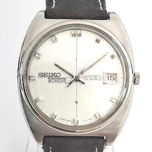 May include: A silver Seiko Lord Matic wristwatch with a white face, black hands, and a black leather strap. The watch face displays the day and date. The watch has a stainless steel case and a black leather strap.