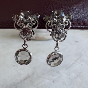 May include: A pair of silver-toned clip-on earrings. Each earring features a large, faceted, clear crystal at the bottom, suspended from a decorative filigree design with smaller crystals. The earrings have a vintage aesthetic.