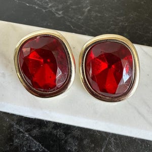 Op de afbeelding: Een paar vintage clip-on oorbellen met grote, facetgeslepen, robijnrode stenen, gezet in goudkleurig metaal. De oorbellen hebben een vierkante vorm met afgeronde hoeken, wat een gedurfde uitstraling creëert. De stenen vangen het licht.