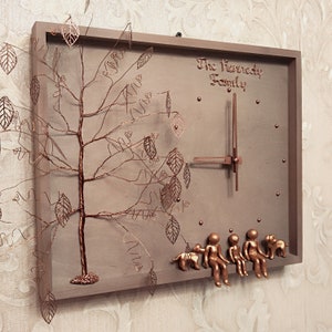 May include: A brown wooden clock with a wire tree design and the text "The Kennedy Family". The clock has a copper-colored face with copper-colored hands and a copper-colored frame. There are five small copper-colored figures sitting on the bottom of the clock.