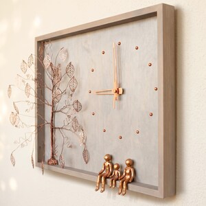 Puede incluir: Un reloj de pared rectangular con marco y esfera de madera gris claro. Presenta manecillas de color cobre, marcadores de hora circulares, un &aacute;rbol tridimensional de alambre de cobre con hojas y tres figuras de color cobre sobre el marco.