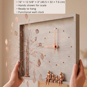 Puede incluir: Un reloj de pared rectangular de madera gris que mide 40,5 cm por 32 cm por 7,6 cm. Presenta un &aacute;rbol de alambre de color cobre con hojas recortadas a la izquierda y cuatro peque&ntilde;as figuras de cobre sentadas en el borde inferior del marco.