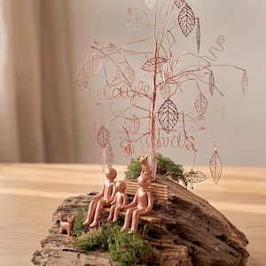 May include: A copper wire sculpture depicting a family seated on a bench beneath a tree, with the names "Caitle" and "Chris" in the branches. A small dog is also present, all on a wooden base with moss.
