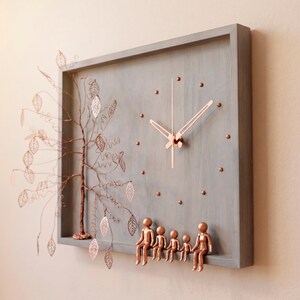 Puede incluir: Un reloj de pared rectangular de madera gris con manecillas y marcadores de hora color cobre. A la izquierda, un &aacute;rbol de alambre de cobre con detalles en forma de hoja sobresale. Cinco figuras color cobre se sientan en el marco inferior.
