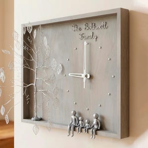 May include: A square, gray wooden wall clock with a silver metal tree and leaf design on the left side. The clock face has the words "The Bothwell Family" and silver dots for the numbers. At the bottom, there are silver figures of a family.