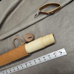 May include: A tan leather tube case with a wooden cap and a matching leather wrist strap. A metal ruler placed alongside shows the case is approximately 17 inches long. The case contains a rolled light-colored paper document or scroll.