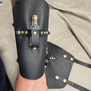 May include: A black leather forearm bracer featuring a small clear glass bottle with a cork stopper secured by a horizontal leather strap and brass-colored metal rivets. The bracer includes adjustable leather straps with matching brass-colored rivets.