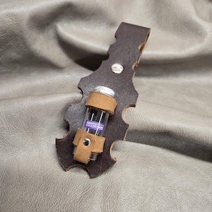 May include: A brown leather vial holder with a decorative shape, holding a small glass vial. The vial has a silver cap and a label with text. The holder is secured with a leather strap and a silver rivet.