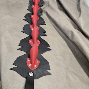 May include: A red and black leather paddle with a twisted design. The paddle features a series of black cut-out shapes with red accents and silver rivets. A black handle is attached at the base.