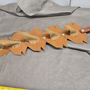 May include: A tan leather strap featuring a series of jagged, leaf-shaped cutouts attached with silver-toned metal rivets. The strap includes a dark brown leather loop at one end and is positioned next to a yellow measuring tape.
