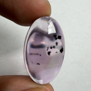May include: Oval-shaped gemstone with a translucent, pale purple hue. The stone features dark, feathery inclusions and is held between fingers. The polished surface reflects light, highlighting the internal patterns and color variations.