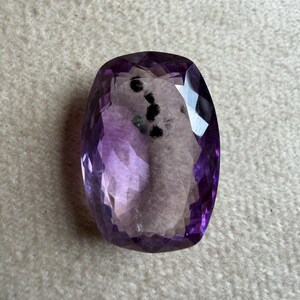 May include: A faceted, oval-shaped amethyst gemstone. The gem is a deep purple color with lighter purple and clear facets. The stone has a polished, reflective surface and is set against a neutral background.
