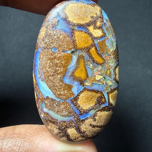 May include: An oval-shaped opal gemstone with a unique pattern. The stone features a mix of colors, including brown, gold, blue, and hints of other colors. The surface has a textured appearance, with a mosaic-like design.