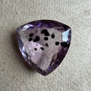 May include: A triangular-shaped gemstone with a faceted surface. The gem is a light purple color with black inclusions. The gem is cut into a triangle shape with a flat bottom and rounded top.