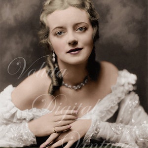May include: A woman with blonde hair styled in braids wears a white lace dress and a pearl necklace. She has her hands clasped in her lap and is looking at the camera.
