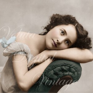 Op de afbeelding: Een jonge vrouw met krullend bruin haar rust haar hoofd op haar arm en kijkt weg. Ze draagt een lichtblauwe en witte jurk met een gerimpelde kraag.