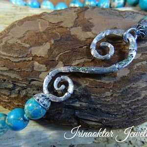 May include: A turquoise blue and green gemstone bracelet with a silver swirl pendant and a silver clasp. The bracelet is on a brown wooden surface.