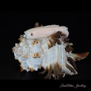 May include: A delicate pink coral fish pendant with a ruby eye and a pearl accent. The pendant is resting on a seashell, suggesting a nautical theme.